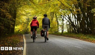 A man and a woman cycle along a rural road with trees on each side.