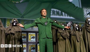 Robert Downey Jr on the Comic Con stage after revealing himself to be playing Doctor Doom. He wears a green suit and his arms are outstretched, with an iron Doctor Doom mask in one hand. He is flanked by several cloaked Doctor Doom lookalikes.