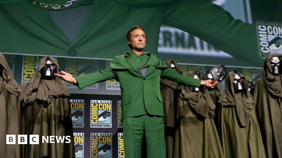Robert Downey Jr on the Comic Con stage after revealing himself to be playing Doctor Doom. He wears a green suit and his arms are outstretched, with an iron Doctor Doom mask in one hand. He is flanked by several cloaked Doctor Doom lookalikes.
