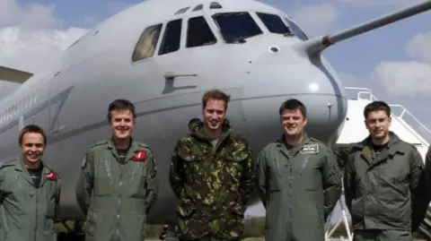 Grant Lawrie Five men are posing in front of a large grey aircraft, on the left is Grant Lawrie, he is smiling in his RAF olive green uniform. In the middle of the group is a young Prince William, he is wearing a camouflage outfit. 