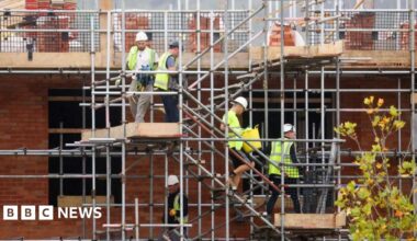 Four men climb down scaffolding stairs on the side of an under-construction brick building. They are wearing yellow high-vis jackets and hard hats.
