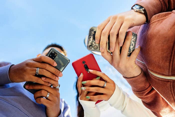 Three people hold smartphones, seen from a low angle under a clear sky, focusing on their devices. Names of individuals are not known