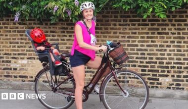 A woman smiling and standing with a bike between her legs. She is wearing a helmet and there is a child on the back with a helmet covering their face.