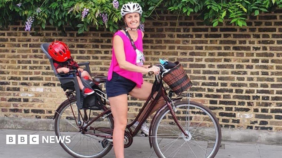 A woman smiling and standing with a bike between her legs. She is wearing a helmet and there is a child on the back with a helmet covering their face.