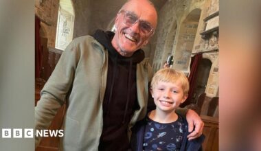 Film director Danny Boyle wearing large framed glasses and a black hoodie in a church with his arm around a young blonde boy wearing a blue dressing gown and a pyjama top with space craft on it.