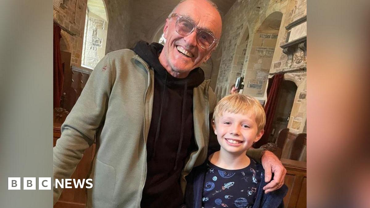 Film director Danny Boyle wearing large framed glasses and a black hoodie in a church with his arm around a young blonde boy wearing a blue dressing gown and a pyjama top with space craft on it.