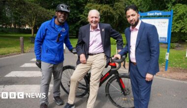 Three men standing in front of a racing bike on a path next to a park. A sign in the background reads East Park. A man at the centre with white hair in beige trousers, pink shirt and blazer leans against it, holding the handlebar. A man on his left is in a blue jacket and cycling helmet, and on his right a man in a navy suit with pink shirt.
