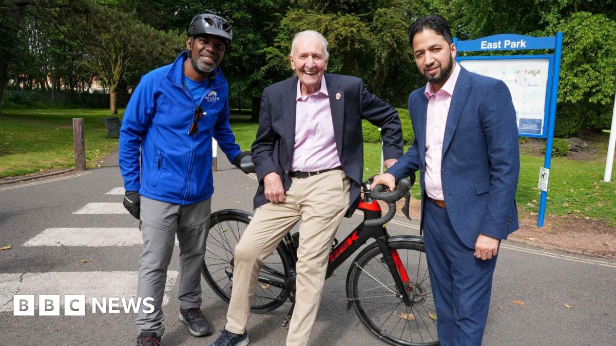 Three men standing in front of a racing bike on a path next to a park. A sign in the background reads East Park. A man at the centre with white hair in beige trousers, pink shirt and blazer leans against it, holding the handlebar. A man on his left is in a blue jacket and cycling helmet, and on his right a man in a navy suit with pink shirt.