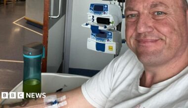 A selfie taken by a man with very short hair wearing a white t-shirt. He is smiling at the camera. Behind him is a small machine and he has an IV line taped onto his arm. Plain walls and floors show he is in a hospital.