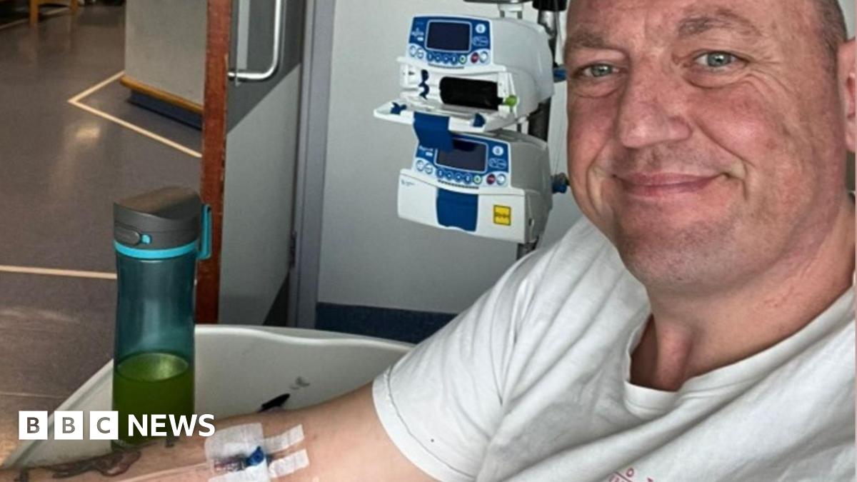 A selfie taken by a man with very short hair wearing a white t-shirt. He is smiling at the camera. Behind him is a small machine and he has an IV line taped onto his arm. Plain walls and floors show he is in a hospital.