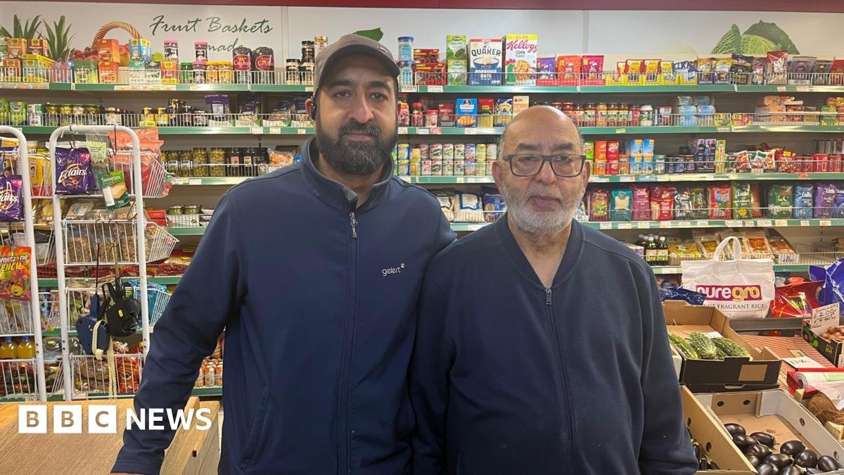 Two men, one on the right in his 60s, the one on the left in his 40s, standing in front of a shop