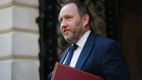 PA Media Ian Murray with dark hair and a greying beard walks in front of a stone building. He is wearing a blue suit and tie with a white shirt, and carrying a red folder. 