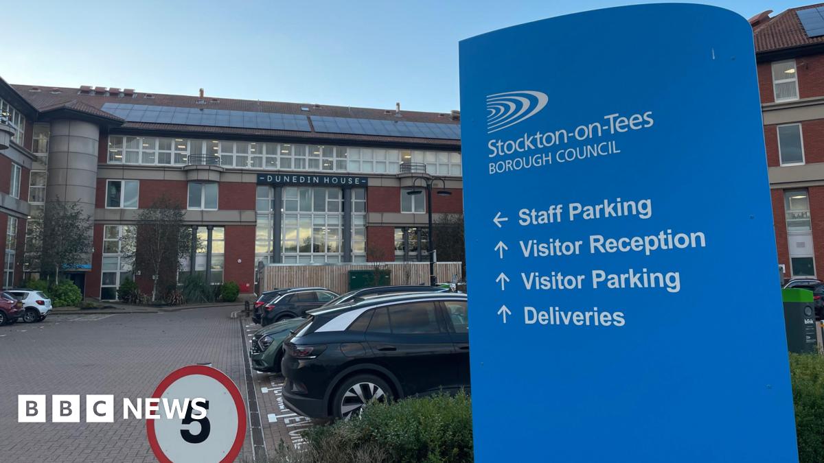 Stockton Council's headquarters Dunedin House. It is a four-storey building mostly made of brown tiles and glass, with solar panels on the roof. A blue sign in the foreground is pointing towards the reception and parking. There are several parked cars and 5mph red and white speed restriction sign.
