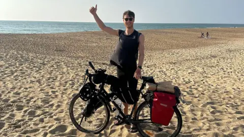 Ollie White Ollie wears a black vest and shorts, and stands with his bike on a very wide sandy beach. The blue ocean can be seen behind him. He has one hand on his bike and the other raised in a thumbs up. 