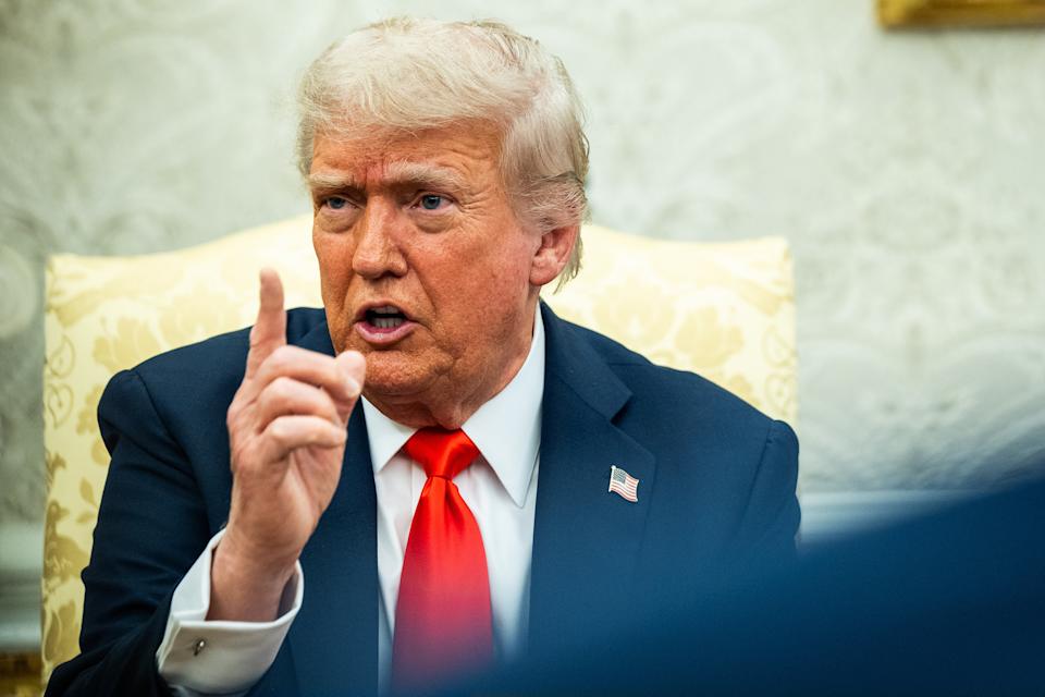 A person in a suit and tie is speaking, pointing a finger for emphasis, seated in an ornate room