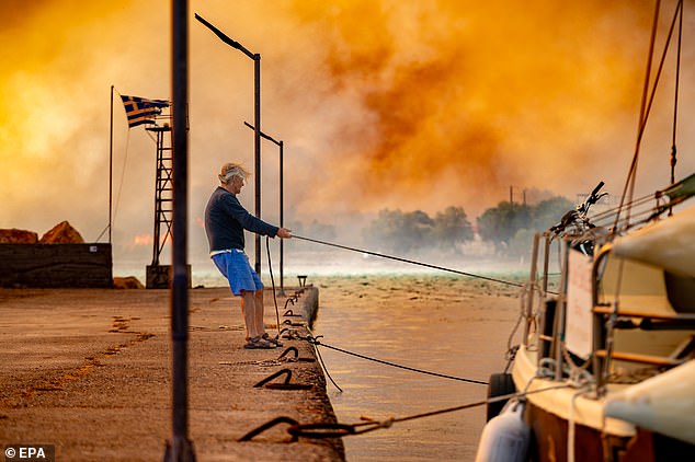 GREECE: Fires have burned throughout the week in Greece. Pictured: A man ties a boat at the port of Lithi in Chios, while a wildfire burns through forested land on June 24