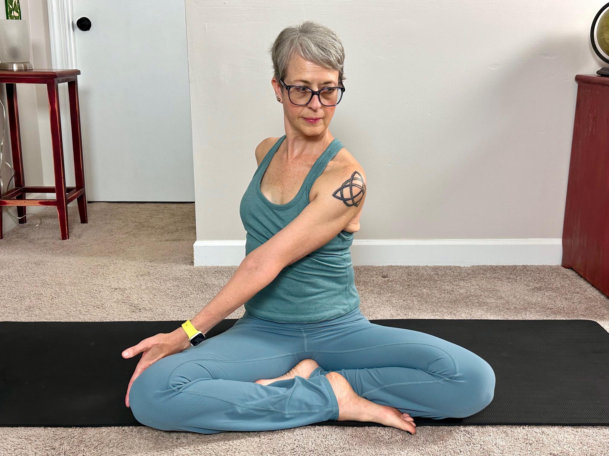 woman sitting cross legged twisting to one side on a black yoga mat in a living room.