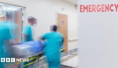 A blurred action shot of three medical professionals wheeling a patient on a trolley down a hospital corridor. The medics ae wearing light blue scrubs. There's a red sign reading 'emergency' on a grey wall.
