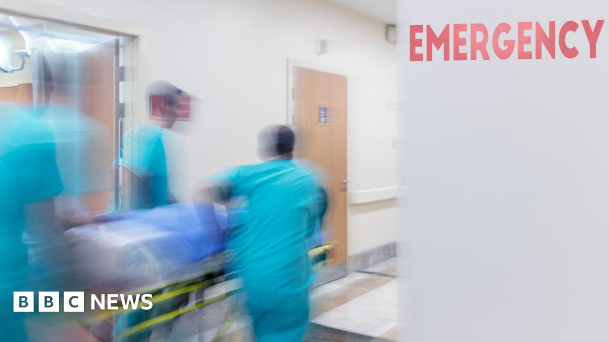A blurred action shot of three medical professionals wheeling a patient on a trolley down a hospital corridor. The medics ae wearing light blue scrubs. There's a red sign reading 'emergency' on a grey wall.