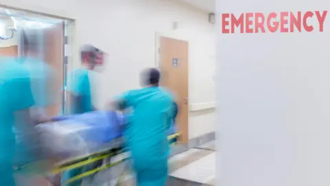 Getty Images A blurred action shot of three medical professionals wheeling a patient on a trolley down a hospital corridor. The medics ae wearing light blue scrubs. There's a red sign reading 'emergency' on a grey wall. 