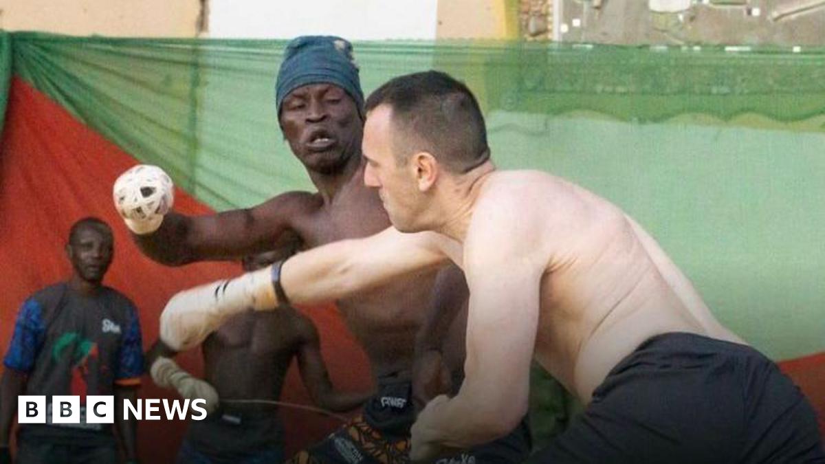 Two topless men, wearing shorts, go to punch one another. Each of one of their fists is wrapped tightly in white rope. There is a Nigerian flag, red and green, behind them on a wall.