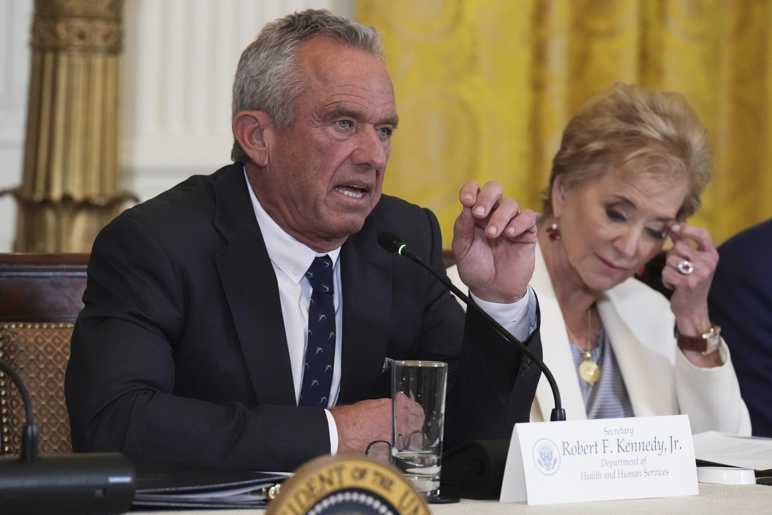 A man in a suit speaks into a microphone at a table with a nameplate; a woman sits beside him, looking down and taking notes.