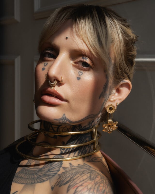 A person with blonde hair, facial piercings, and multiple tattoos poses in sunlight. They wear gold jewelry, including hoop earrings, a nose ring, and stacked neck rings, with visible tattoos on their face, neck, and chest.