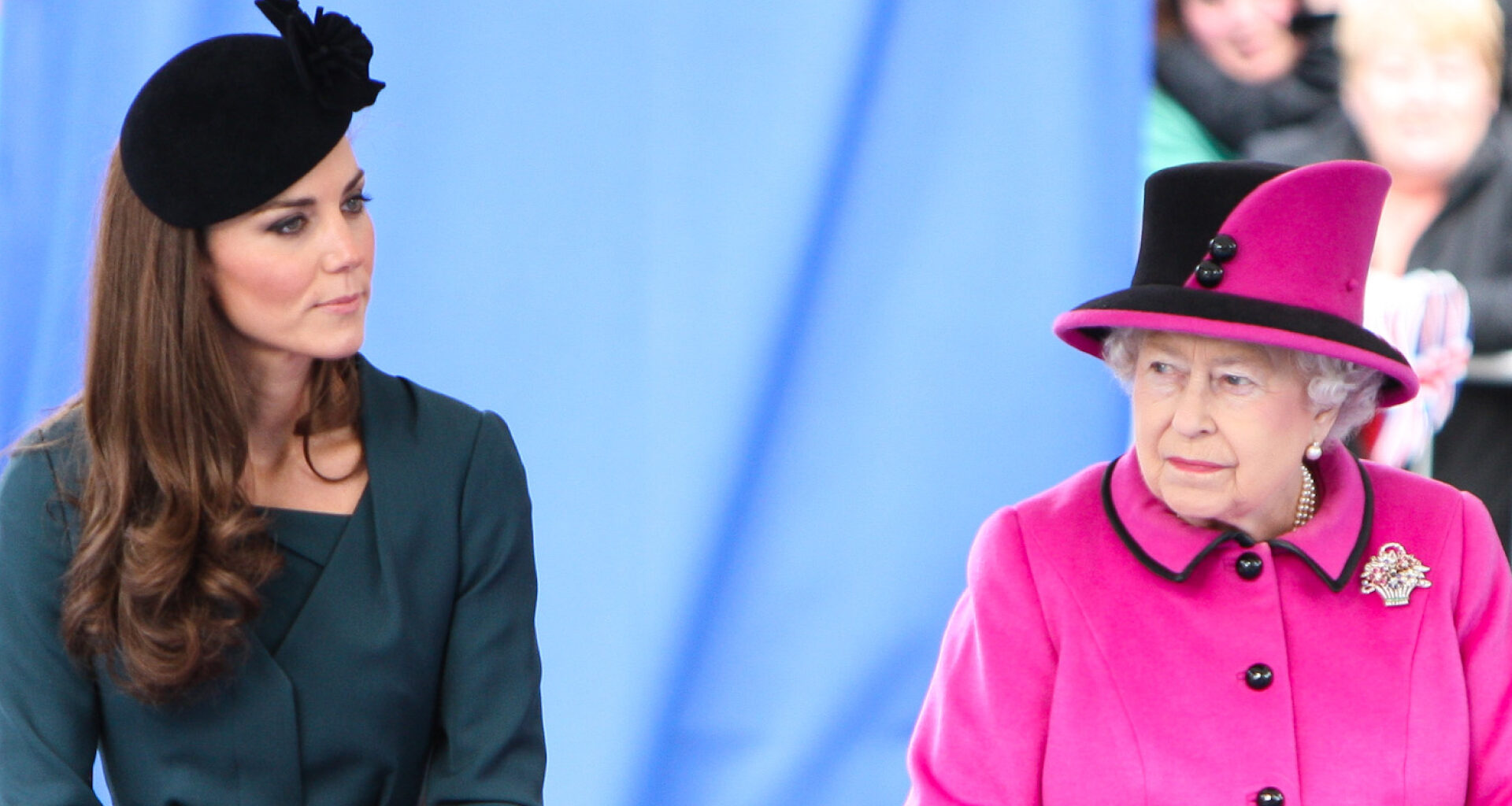 Kate Middleton wears a dark green dress with a black hat and Queen Elizabeth wears a bright pink coat