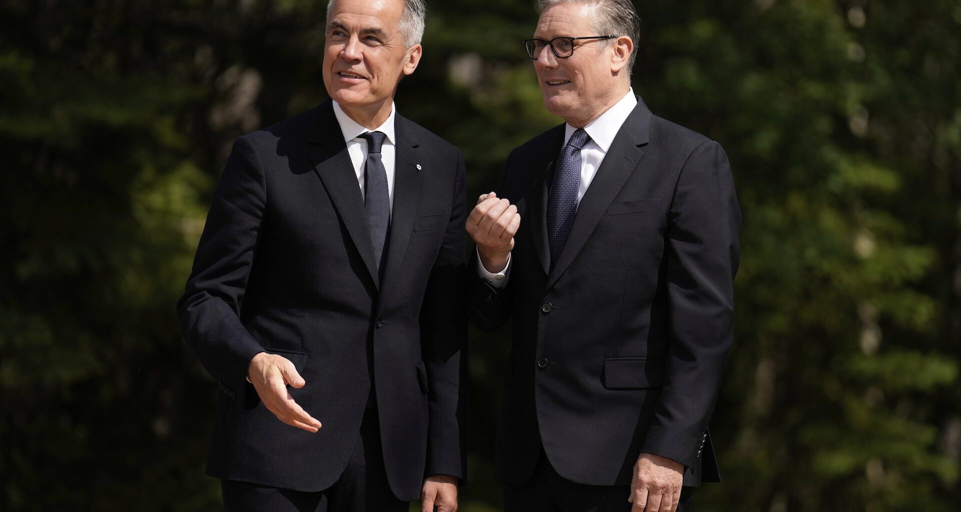 A shot of Mark Carney and Keir Starmer against a backdrop of evergreen trees. Both are smiling and look at something in the middle distance.