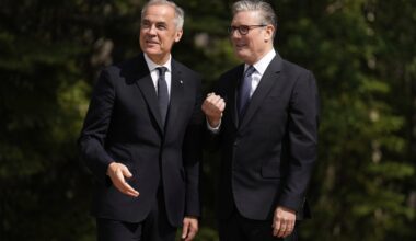 A shot of Mark Carney and Keir Starmer against a backdrop of evergreen trees. Both are smiling and look at something in the middle distance.