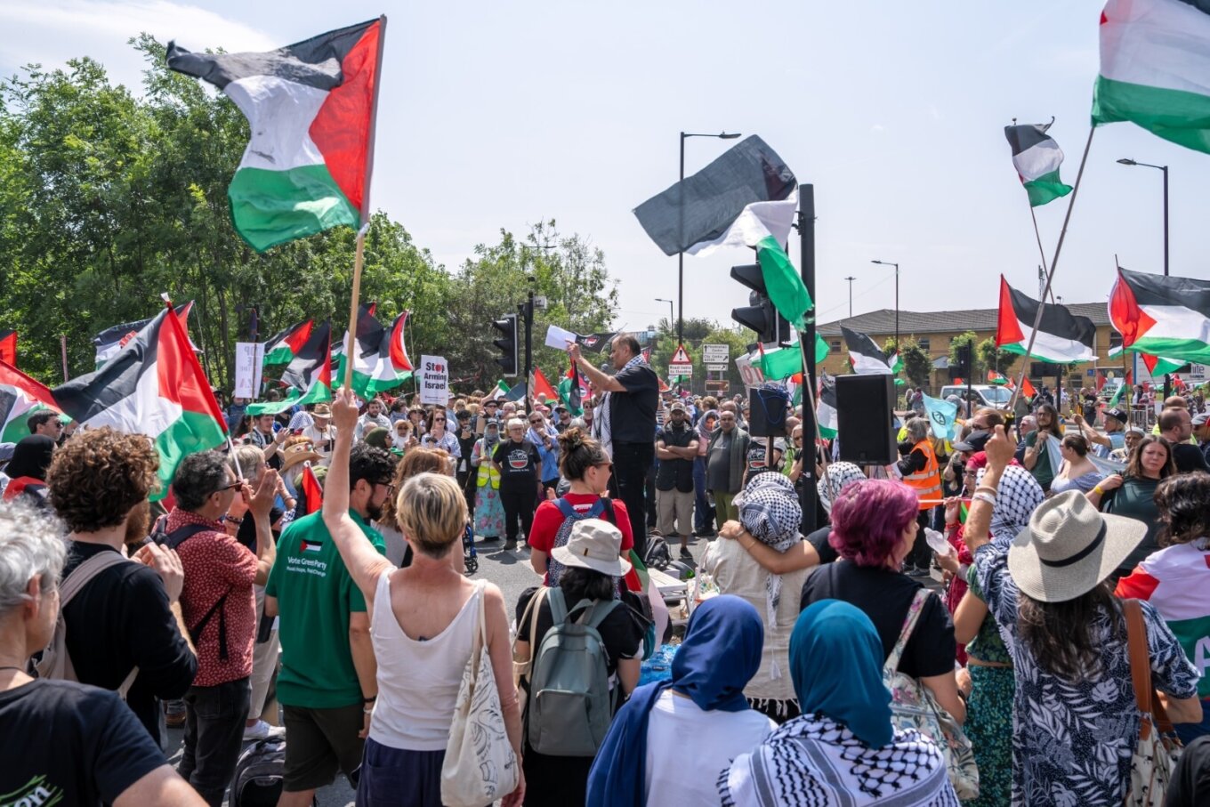 A photo of a crowd of a few hundred people at a protest, holding Palestinian flags and listening to a speaker