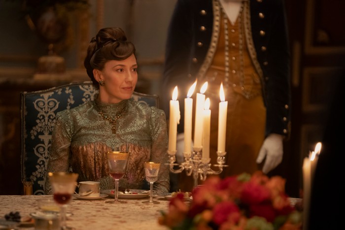 Carrie Coon in The Gilded Age, wearing period dress and sat at a candlelit dinner table.