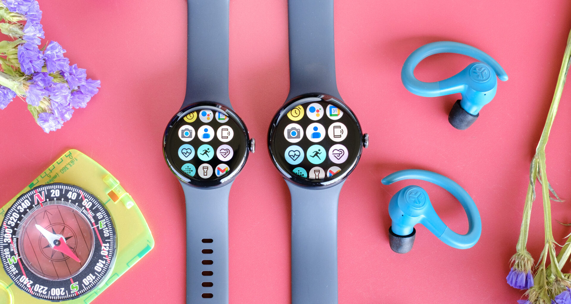 The Google Pixel Watch 3 in 41mm and 45mm sitting beside each other on a red table next to blue earbuds, purple flowers and a yellow compass