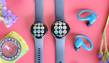 The Google Pixel Watch 3 in 41mm and 45mm sitting beside each other on a red table next to blue earbuds, purple flowers and a yellow compass