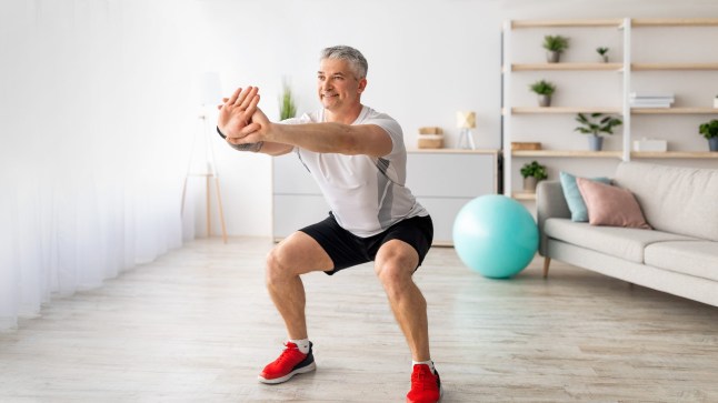 Strength workout concept. Active mature man doing squats at home, exercising in living room, panorama, free space