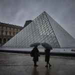 Two people shelter under an umbrella during a rainy day next to a glass pyramid structure.