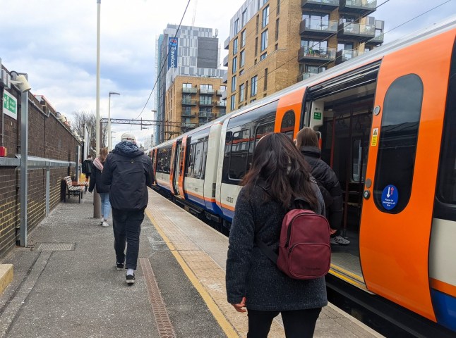 Diverse commuters walk past a London Overground train at the Walthamstow Central station platform