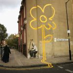 A woman walks past a some a painting of a yellow flower on a wall on a street.