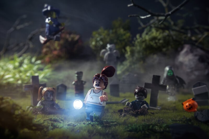 A LEGO figure holding a flashlight stands in a foggy, spooky graveyard surrounded by tombstones, creepy figures, and ghosts, with a witch flying on a broom in the background and a pumpkin on the ground.
