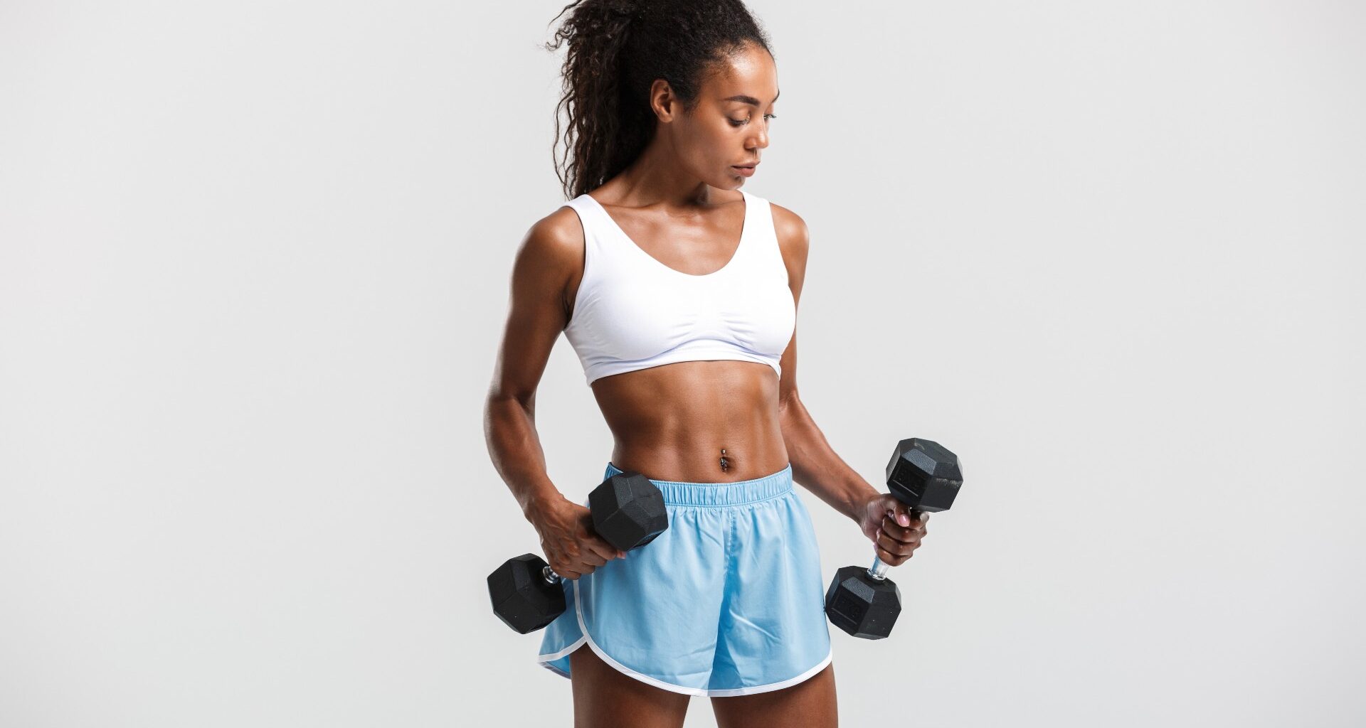 Woman against a grey backdrop in workout clothes holding two dumbbells in front of her during dumbbell workout