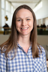 A woman in a blue plaid shirt smiles at the camera, the TMI in the background