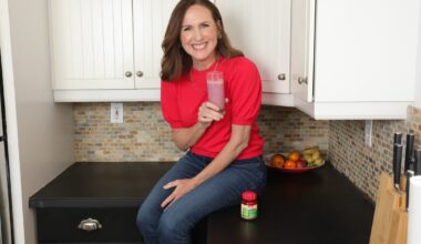 Molly Shannon sitting on a black marble countertop in a kitchen holding a pink smoothie.