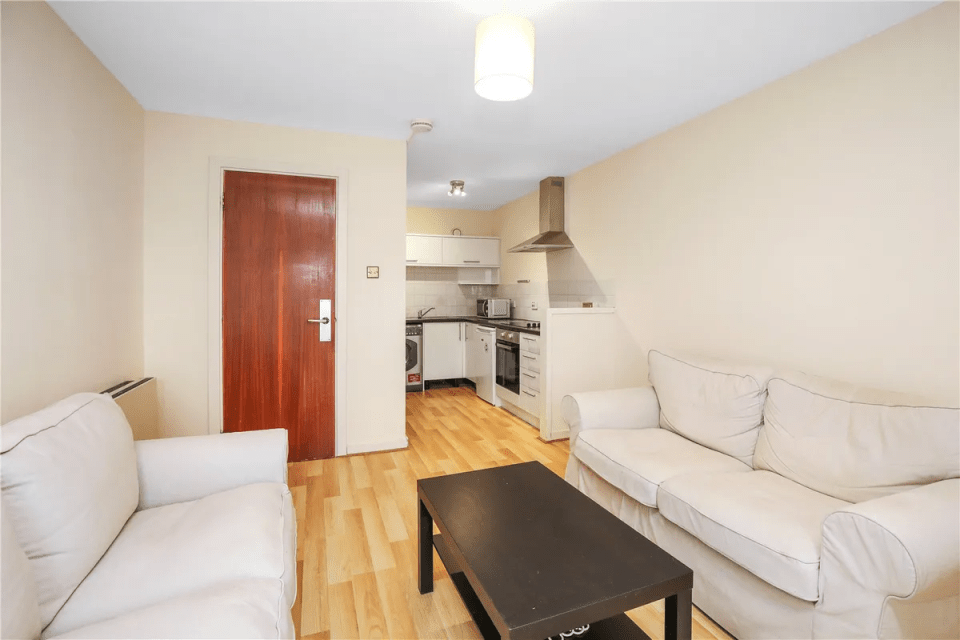 Apartment living room with off-white sofas and a view of a small kitchen.