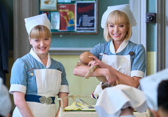 In a scene from Call the Midwife, two characters wear blue nurse uniforms and smile. One holds a toy baby.