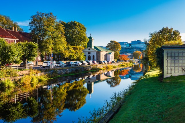 The calm water of a canal reflects a church building in Gothenburg, Sweden. The setting is idyllic with bright blue skies, green trees, and a row of parked cars lining the bank. A lush, green lawn slopes down to the water's edge.