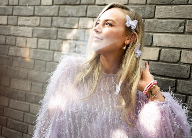 Georgie Swallow in a lilac glitzy dress stood in front of a brick wall. She is looking off into the distance with a hand touching her hair. She has butterfly clips in her blonde hair