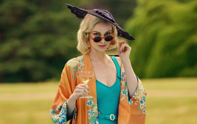 Joanna Vanderham as Diana Mitford, wearing a purple wide-brimmed hat, orange silk kimono and blue swimsuit while holding a glass of champagne.