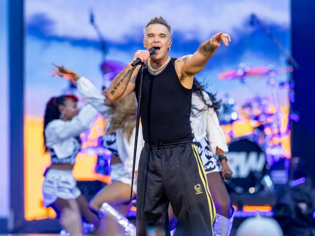 Robbie Williams sings in a black vest and trackies, with dancers performing behind him.