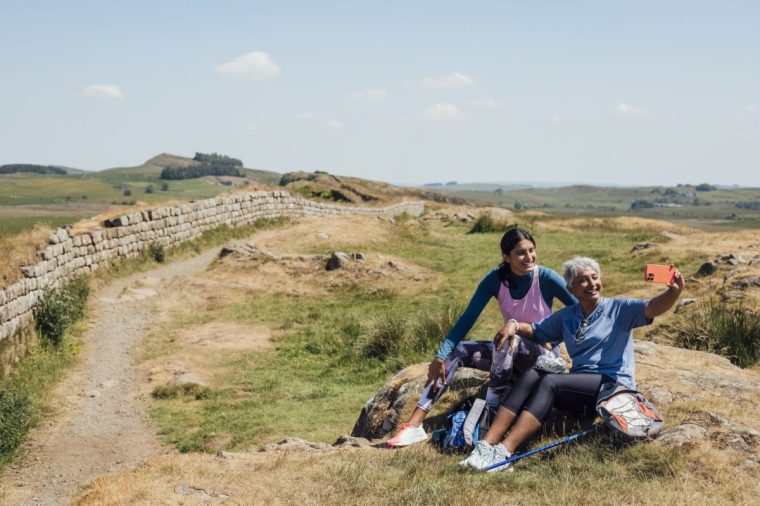 A full shot of Mother and Daughter sitting on a rock in the countryside, they are happily smiling whilst the mother holds up her phone to capture a photo. They're both wearing casual sports attire on a summer's day at Hadrian's Wall, Northumberland. Videos are available similar to this scenario.