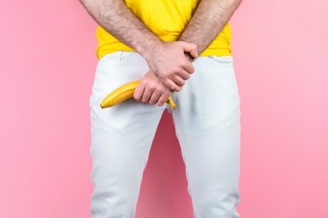 Potency and men's health. A man in white jeans, legs apart, holds a banana near the genitals. Pink background. Close up of hands.